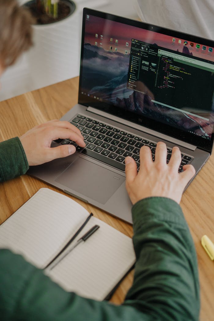 our-story A person coding on a laptop with a notebook at a wooden desk in a modern indoor setting.